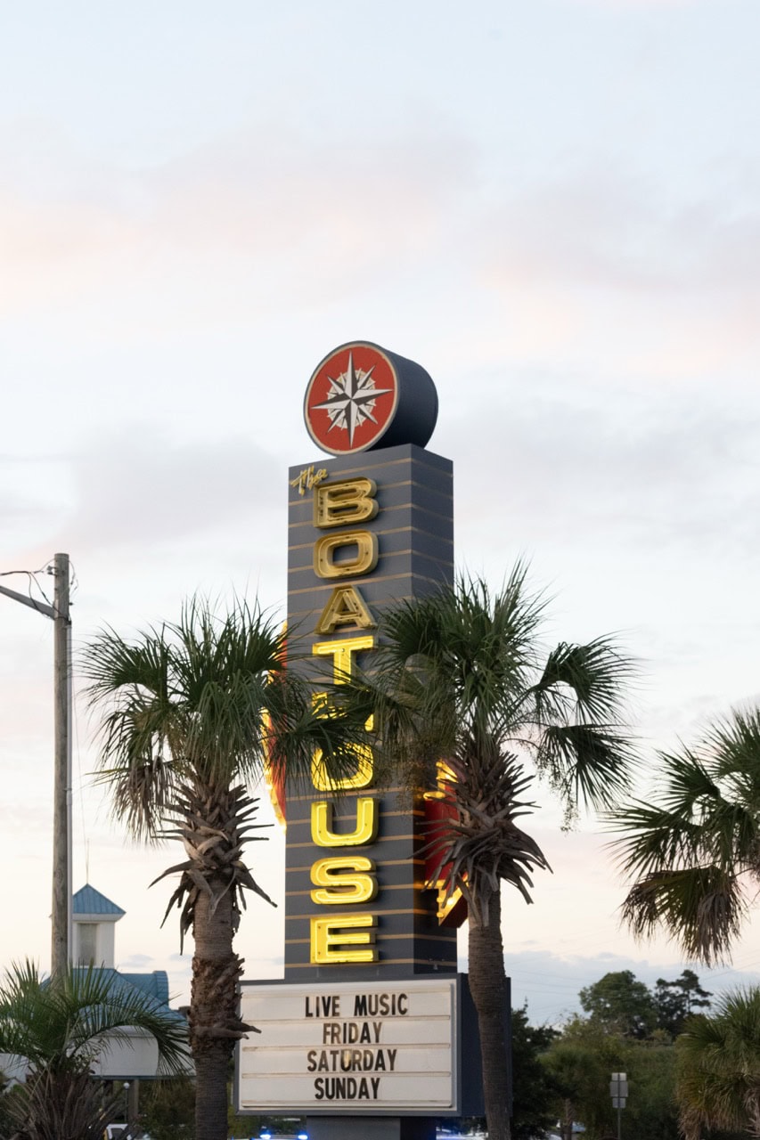 Mustang Week sign at Myrtle Beach with palm trees and event details.