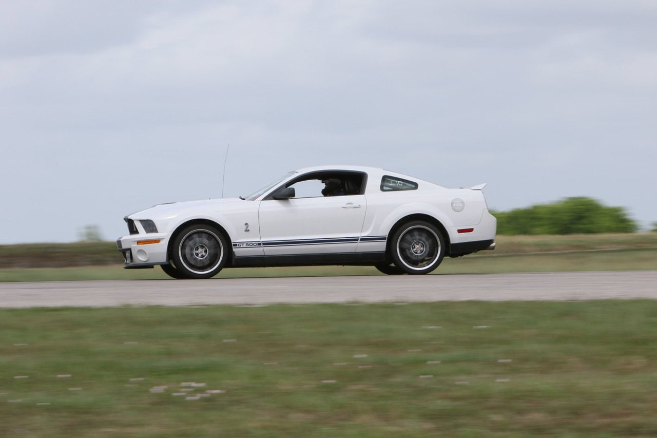 Classic white Ford Mustang driving on open road during Mustang Week event.