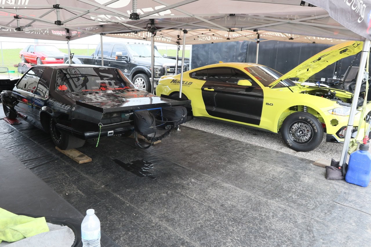Two Ford Mustangs at Mustang Week car show, one black and one yellow, under a canopy.