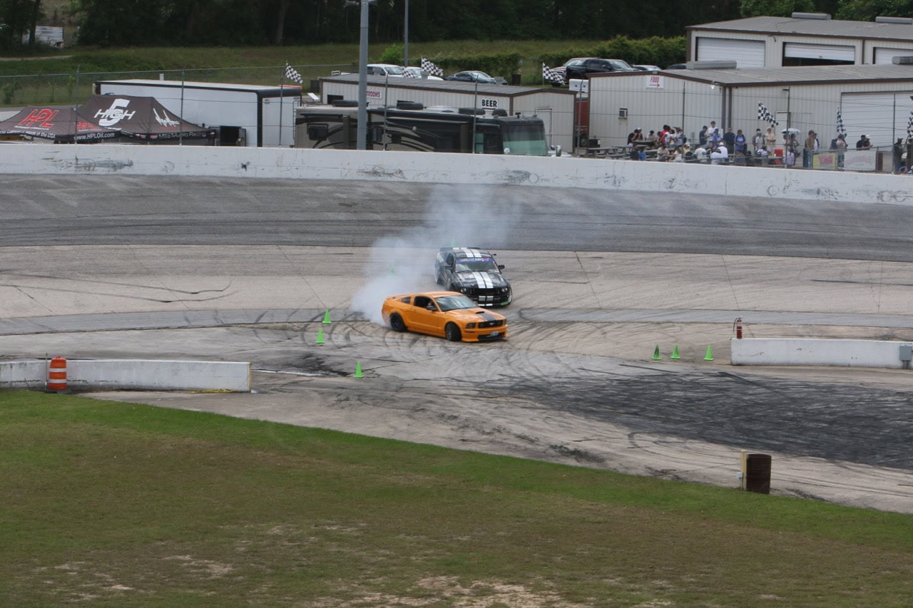 Mustang Week drifting event with orange and black cars on the track.