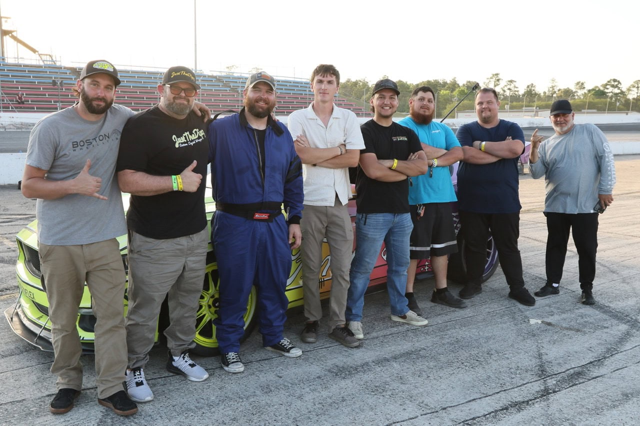 Group of men standing next to a race car at Mustang Week event.