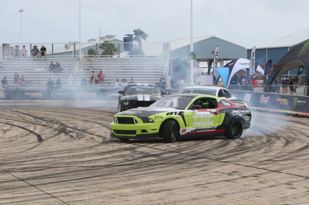 Mustang Week drift car performing a stunt on the track with tire smoke and spectators in the backgro.