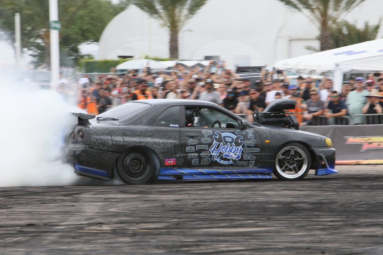 High-performance Mustang drifting at Mustang Week event with crowd and palm trees in background.