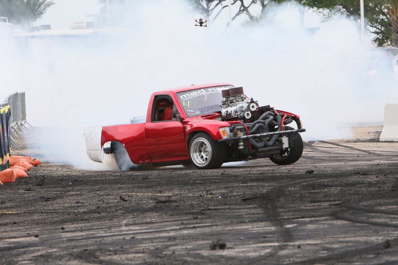 Red pickup truck performing burnout at Mustang Week drag race event.