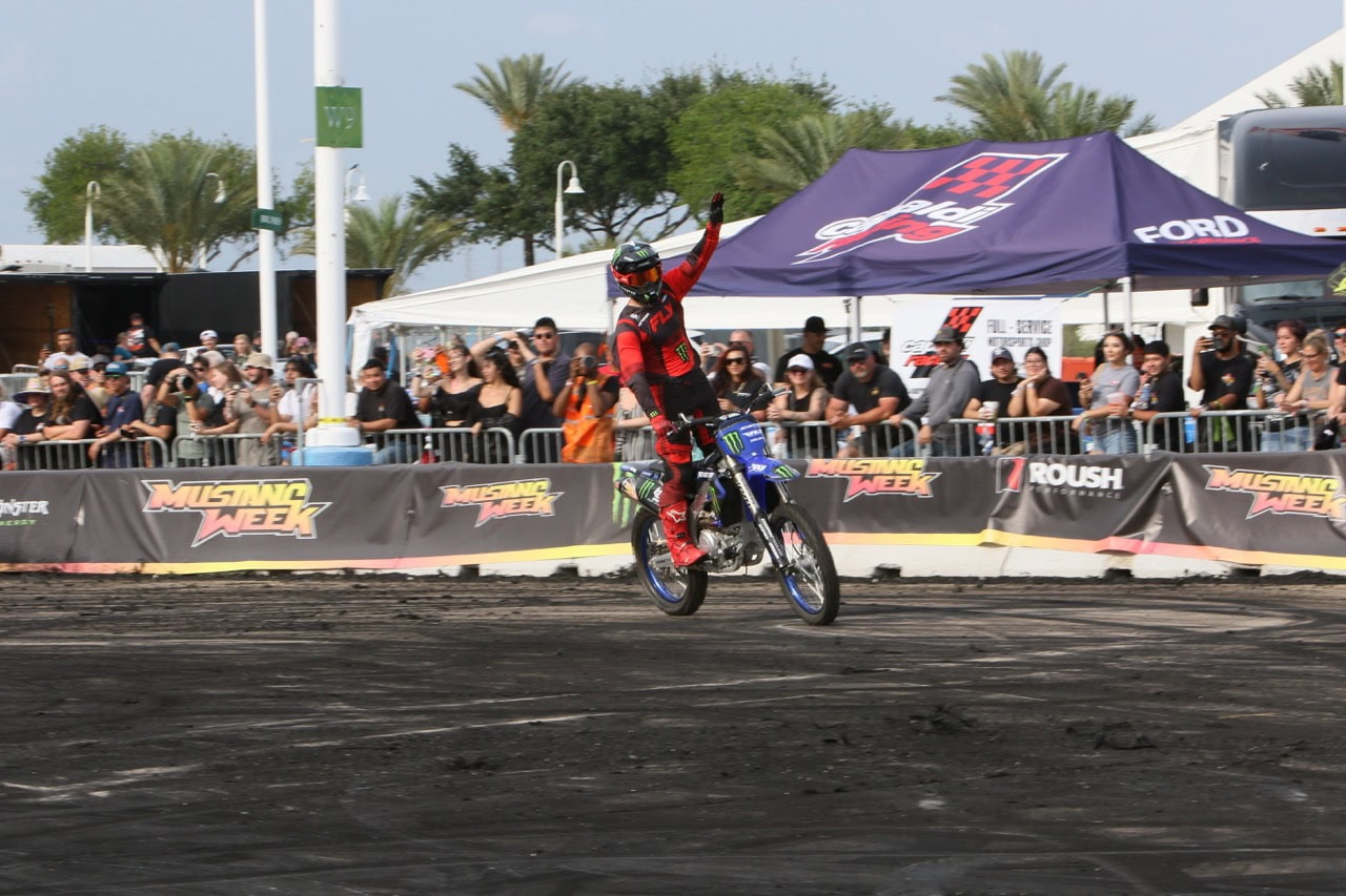 Dirt bike stunt rider performing wheelie at Mustang Week event with crowd and tents in background.