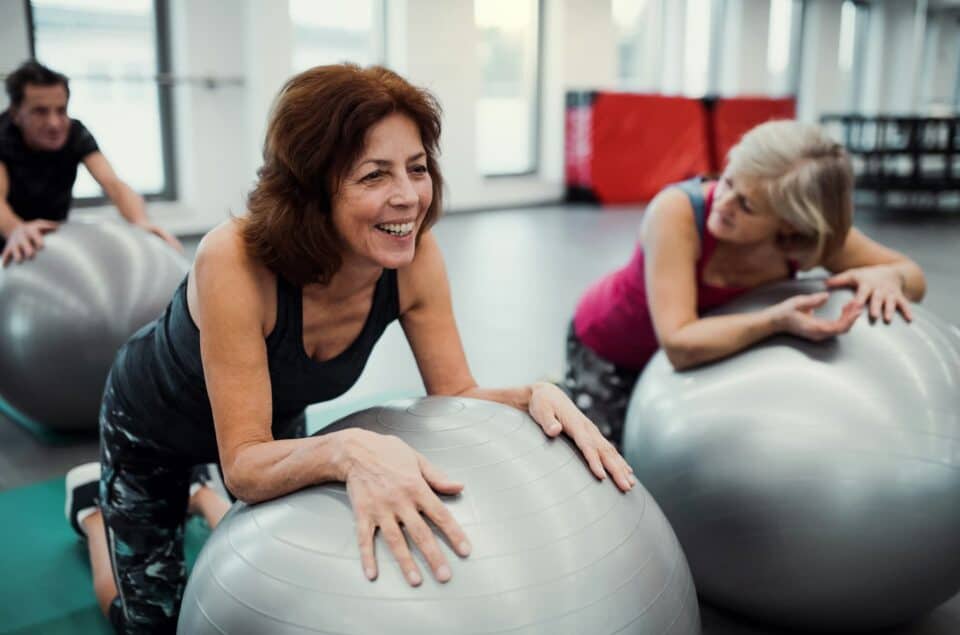 3 older adults attending an excercise class. 2 women up front are resting on inflattable exercise ball. They are talking to each other
