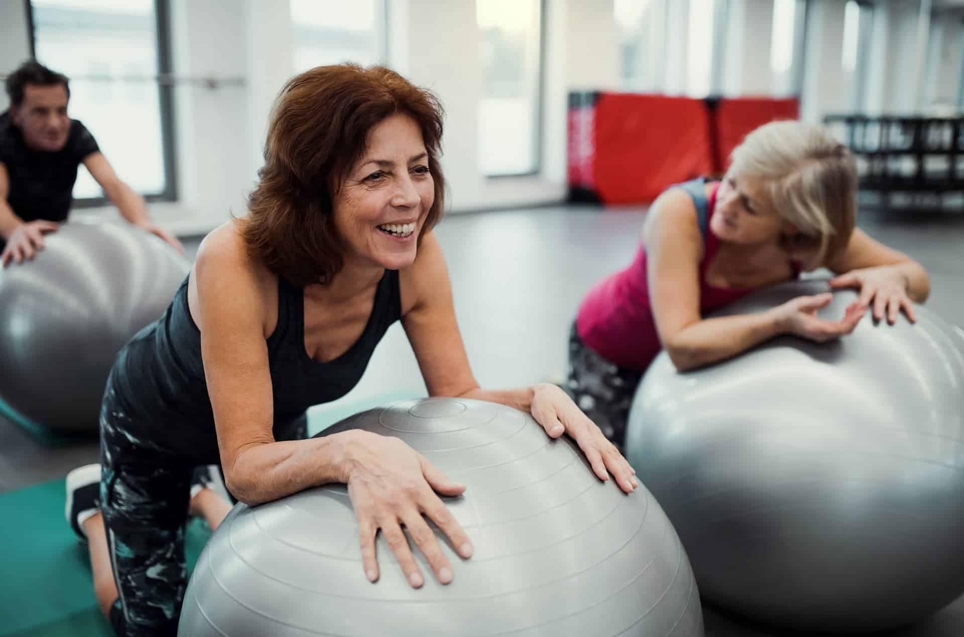 3 older adults attending an excercise class. 2 women up front are resting on inflattable exercise ball. They are talking to each other