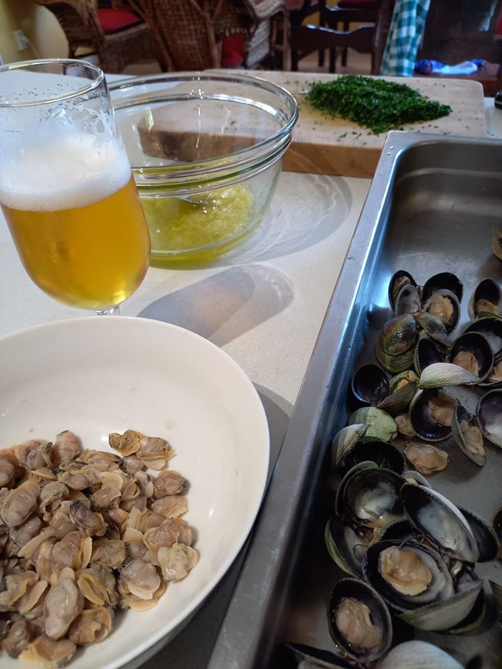 De shelling clams. A oven tray with steamed clams, a white bowl with clam meat, a beer in a glass and a clear bowl . A chopping board with cut herbs in the background