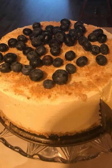 A cheesecake topped with blueberries on a glass platter standing on a white table. A cake serving cutter slicing into the cake