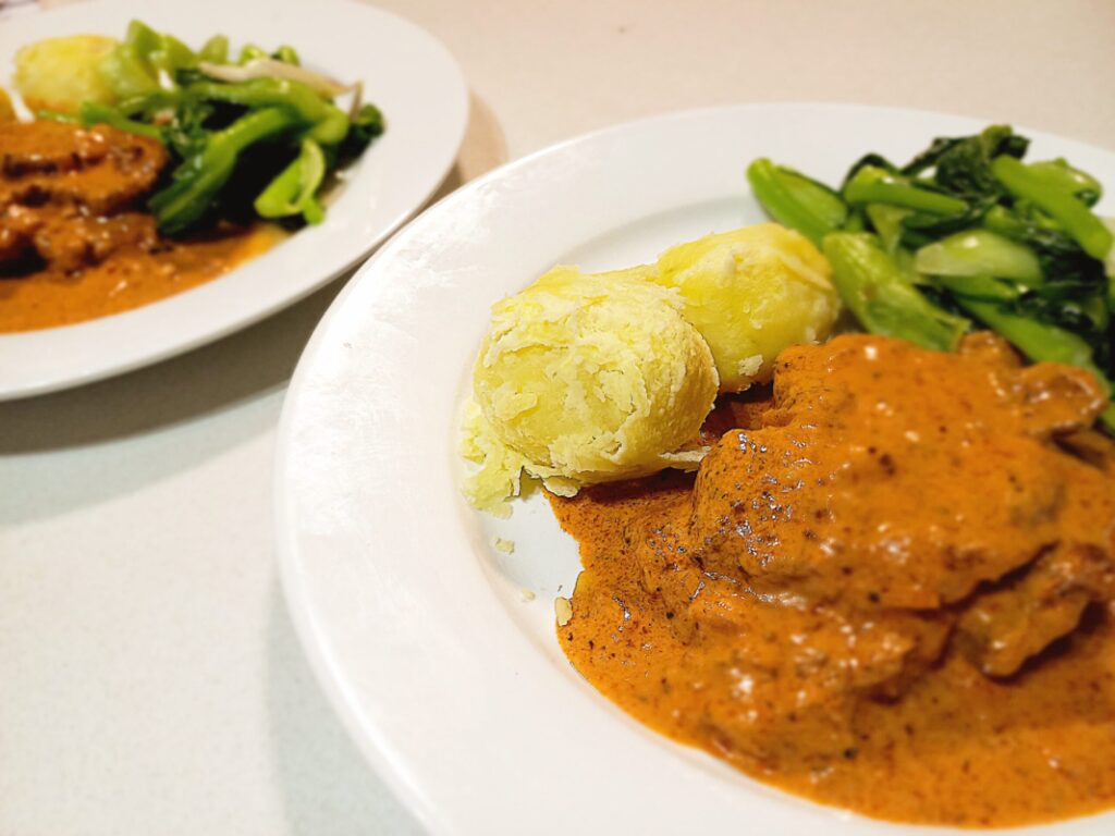 two plates of pork pepper steak in a tomato cream sauce with boild potatoes and steamed vegetables on a white kitchen bench