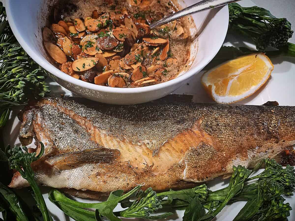 Whole trout, a white bowl with almondine sauce, steamed broccolini and a lemon wedge served on a white plate.