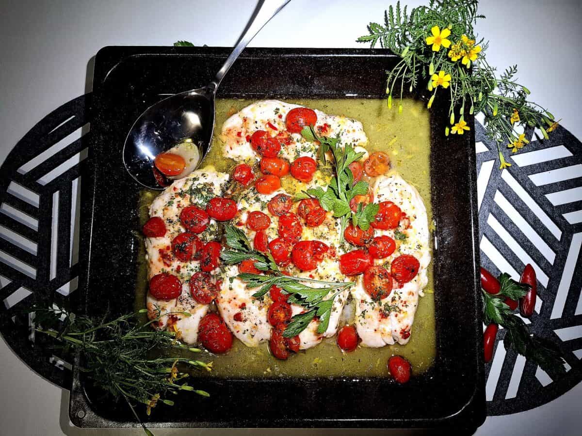 4 Baked fish fillets in an oven tray topped with tomato garlic and parsley. On a round place mat on a white table. Silver serving spoon, garnished with yellow flowers.