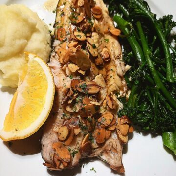A Trout fillet on a white plate topped with almondine sauce. Mash potato and a lemon wedge on one side and steamed broccolini on the other side of the fillet