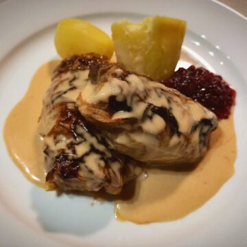Two Swedish cabbage rolls on a white plate with two boiled potatoes, creamy sauce and lingon berry jam