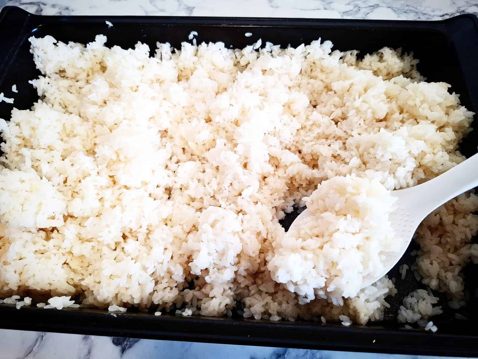 Sushi rice seasoned with sushi vinegar in a black oven tray. A white rice spoon holding up a spoon of rice