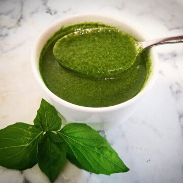 Basil garlic oil in a white ramekin with a spoon holding up some of the oil. Fresh basil leaves in front of it