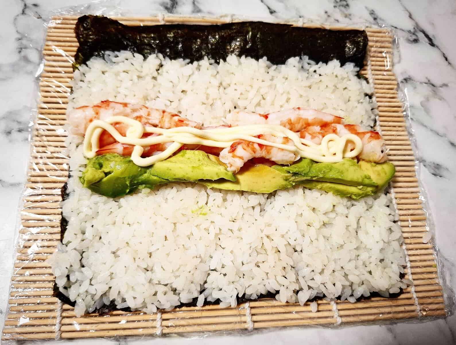 Sushi rice spread out on top of a nori sheet on a sushi mat covered with cling wrap on a marbled bench top. Cooked peeled prawns and slices of avocado are laid out in a horizontal line on top of the rice in the middle with Kewpie mayonnaise drizzled on top of it.