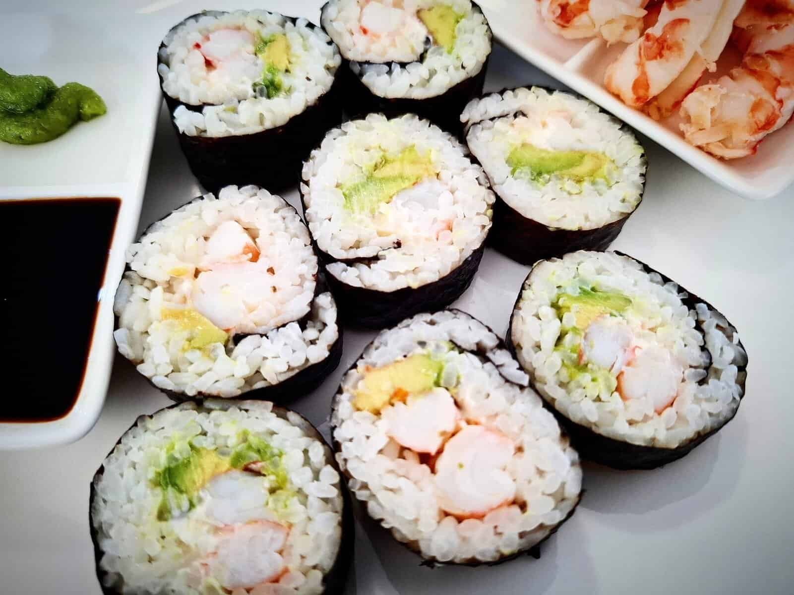 Eight pieces of prawn and avocado sushi on a white plate with a few peeled prawns next to it on the right hand side and a double ramekin of soy sauce and wasabi on the left hand side of the sushi pieces