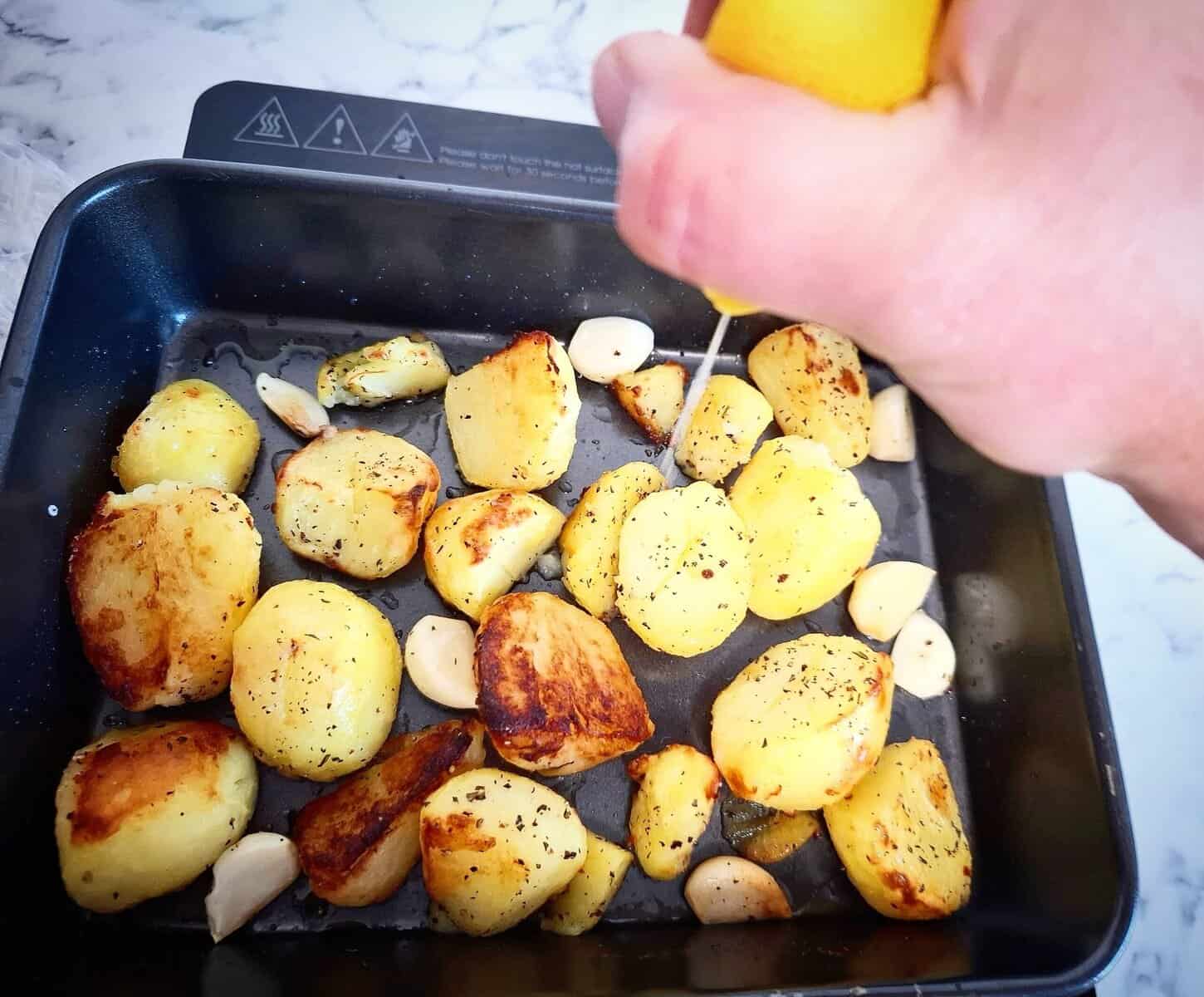 Adding garlic cloves and lemon juice to the roasted potatoes in a black oven tray