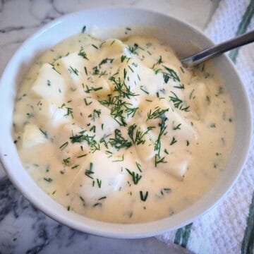 Dill creamed potatoes in a white bowl and a serving spoon in it. The bowl is placed on a marbled bench top with a white and green stripe towel next to it.