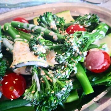 Steamed broccolini, green beans, zucchini slices and cherry tomatoes on an oval silver plate with tahini garlic dressing drizzled over the top garnished with sesame seeds. A white and green tea towel next to it.