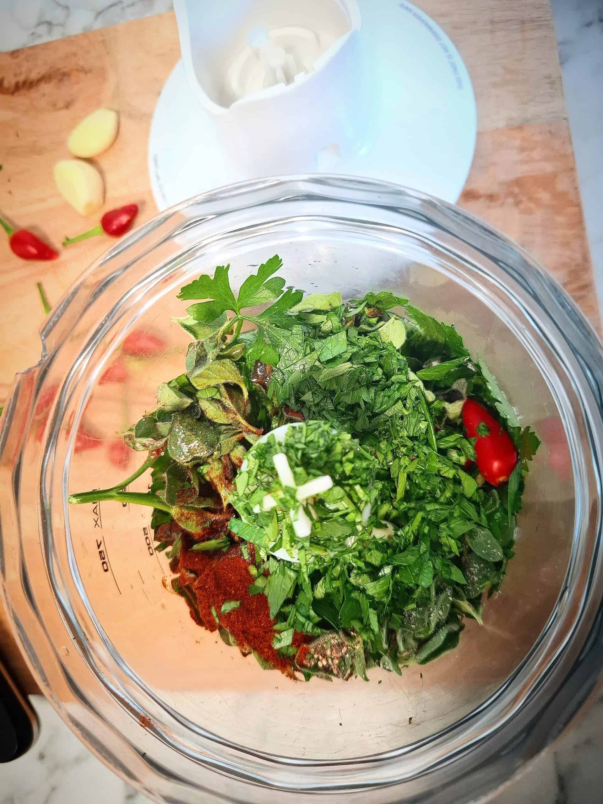 Ingredients for chimichurri sauce red in a clear blender base ready to be chopped. The blender base and the white lid is on a wooden chopping board with red chili' and peeled garlic cloves on it.