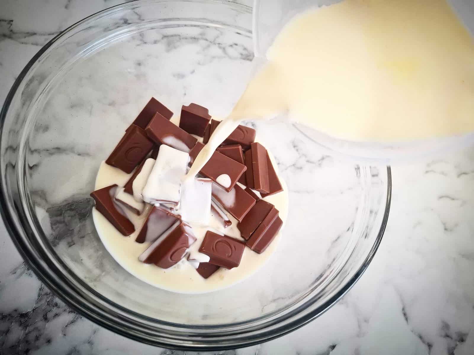 Pouring hot cream from a plastic jug over rectangular chocolate pieces in a glass bowl on a marbled bench top