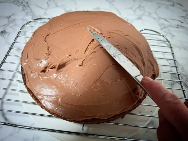 Spreading chocolate ganache on top of the banana cake with a butter knife while it's on a wired wrack.