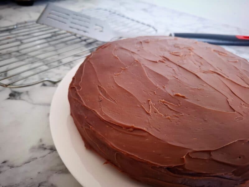 An iced banana chocolate cake on a round white plate. A wired wrack with a large spatula on top behind it.
