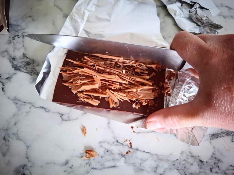 Making chocolate shavings by scraping a sharp knife along the bottom flat side of a chocolate bar with foil wrapping underneath it.
