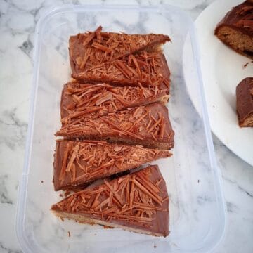 Six pieces of banana cake with chocolate icing in a rectangular plastic tub on a marbled bench top. The corner of a round, white plate with the rest of the cake next to it.