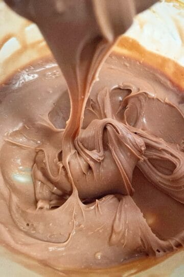 Chocolate ganache has cooled down to a thick but spreadable consistency good for icing a cake. The ganache is in a glass bowl and a spoon holding some up above it with some ganache slowly flowing down into the bowl.