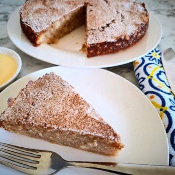 Cooked apple and almond cake dusted with cinnamon and icing sugar on a white round plate on a marbled bench top. A wedge portion has been cut from the cake and served on a white plate with a fork. A White, blue and yellow napkin with cake serving cutlery on top next to it and a white ramekin with custard in it on the other side next to the cake.