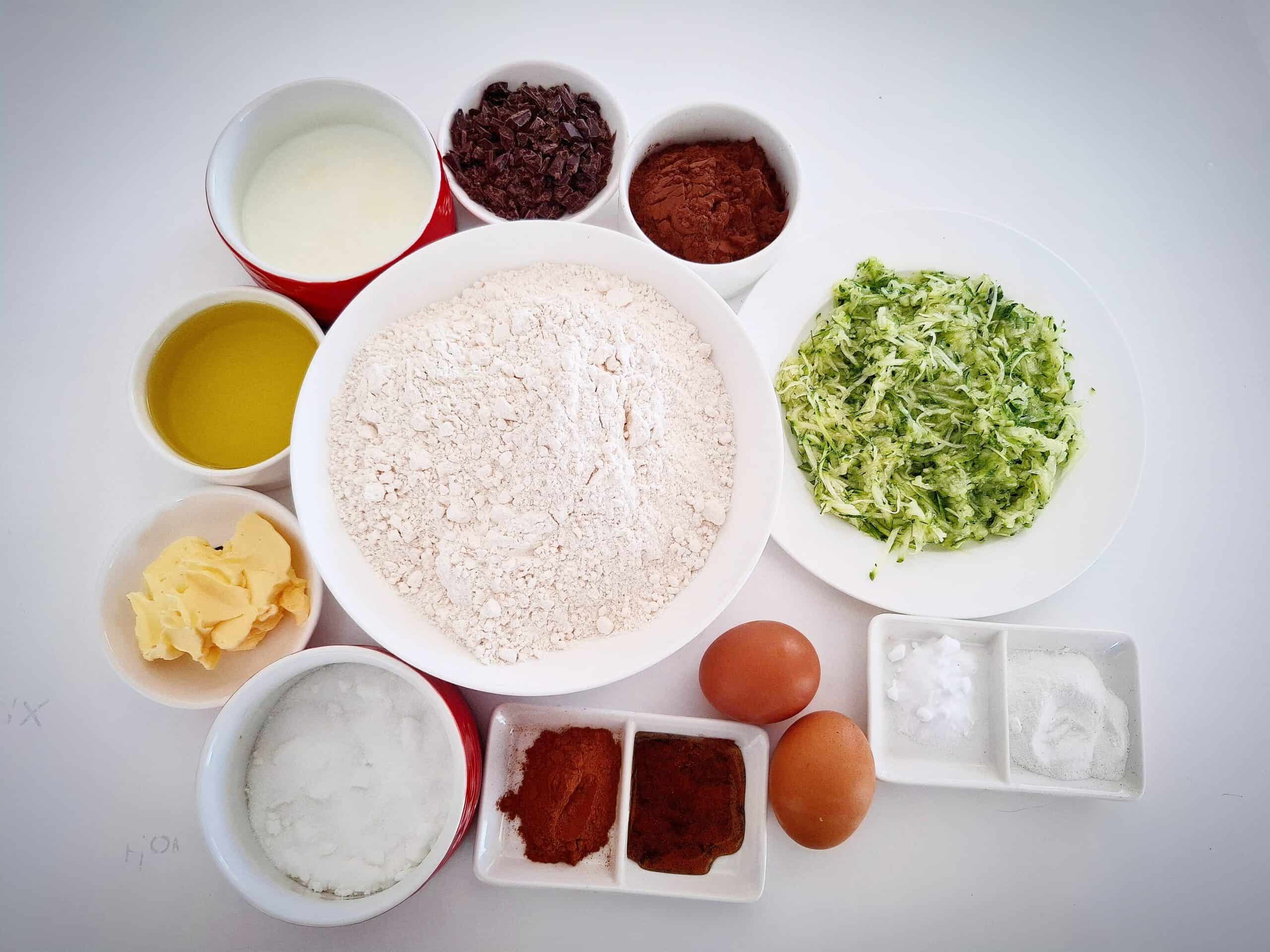 Ingredients for courgette and chocolate cake in ramekins on a white table.