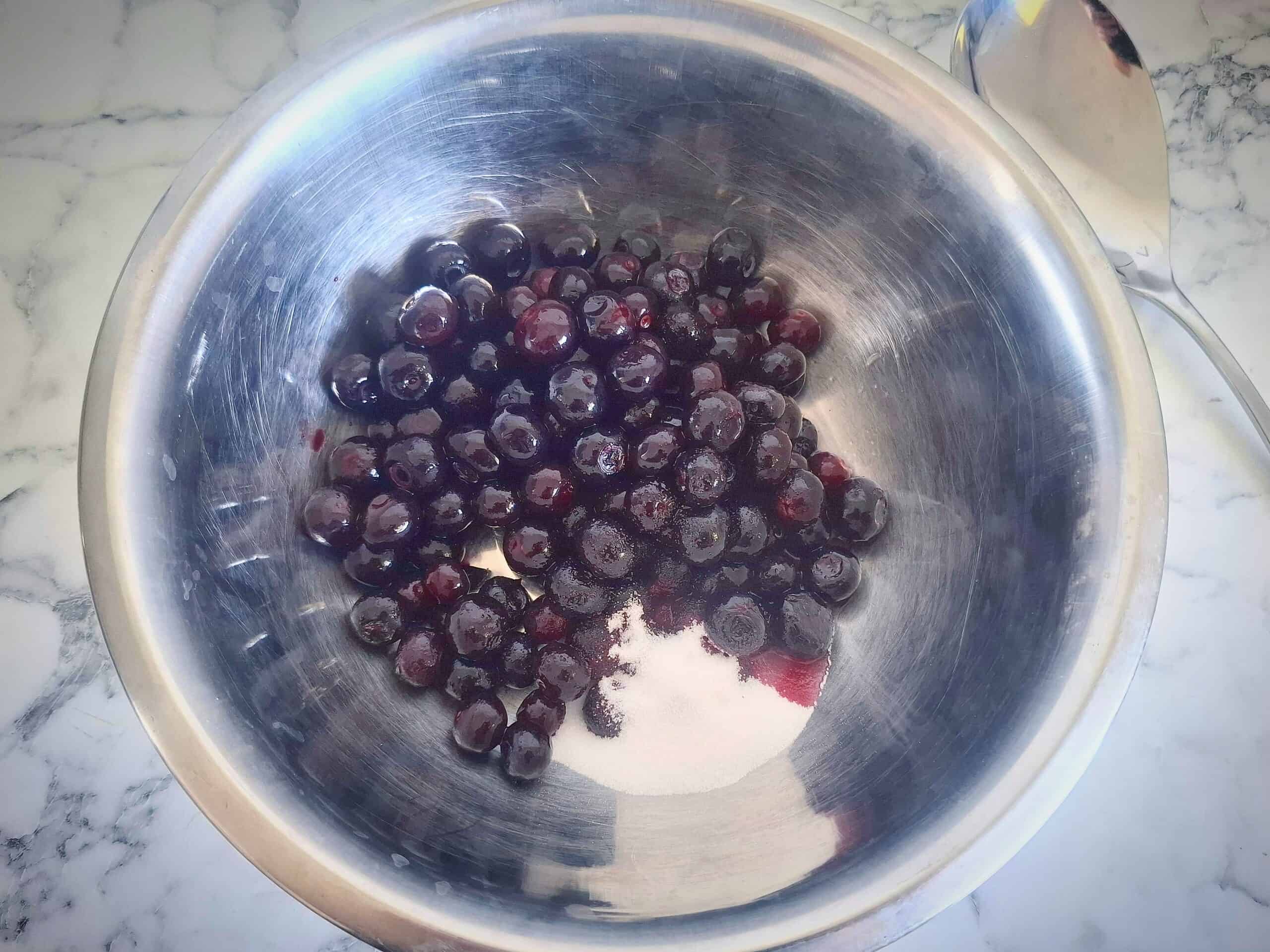 Blueberries, sugar and lemon juice in a stainless steel bowl. A silver spoon next to it.