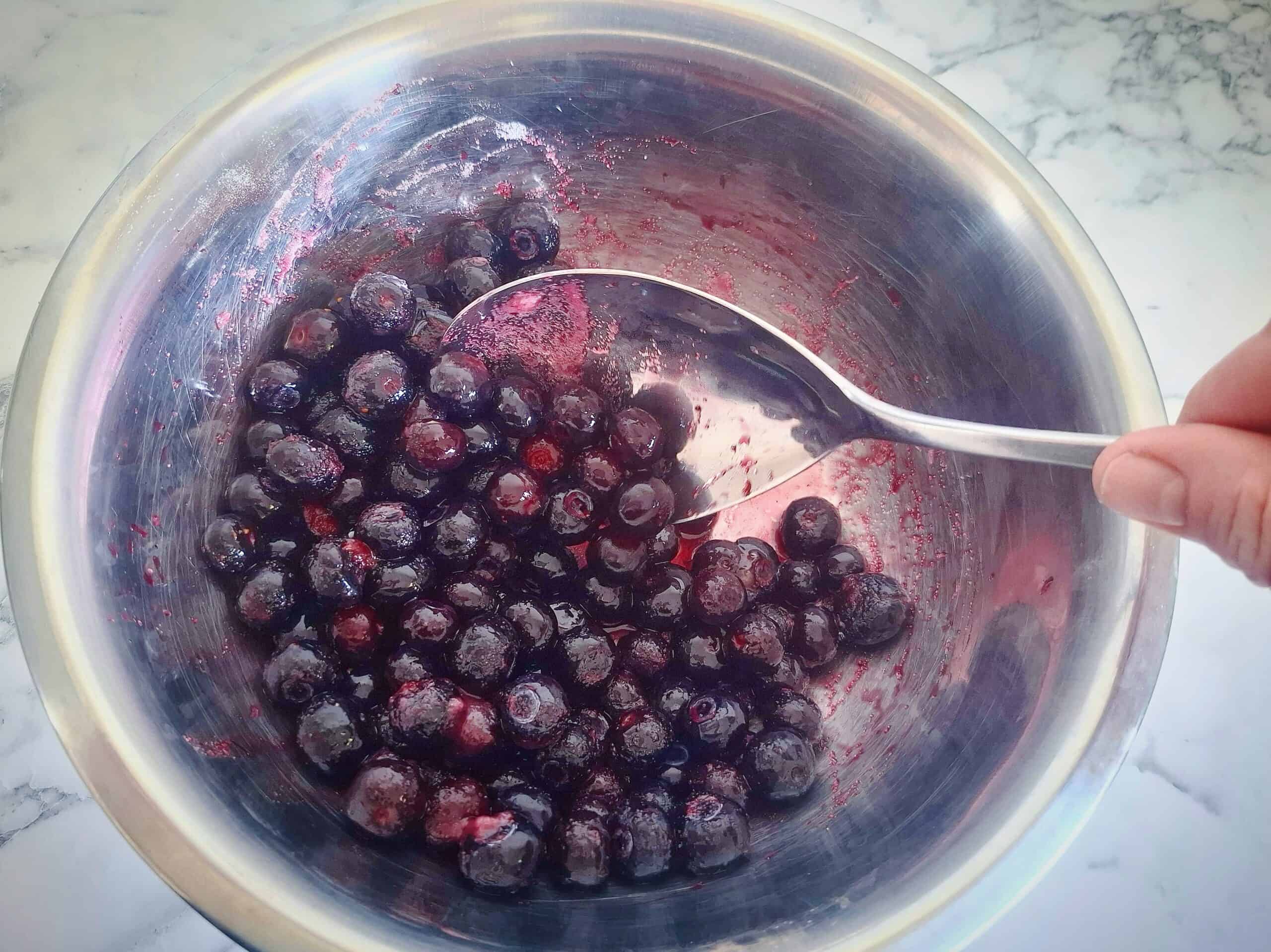 Mixing blueberries with sugar and lemon juice with a silver spoon in a stainless steel bowl