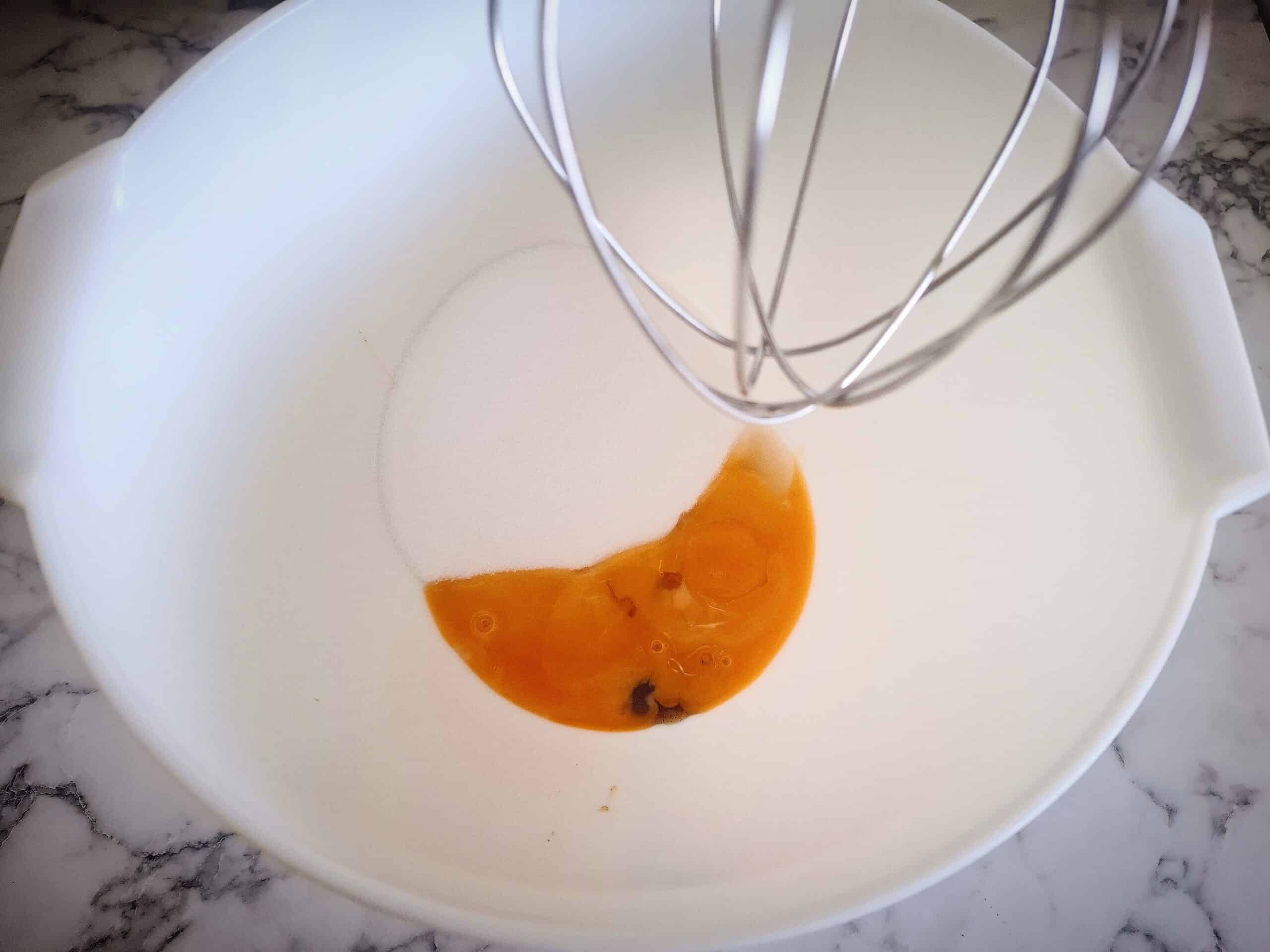 Egg yolks, sugar and vanilla in a white bowl. A kitchen mixer whisk above it.