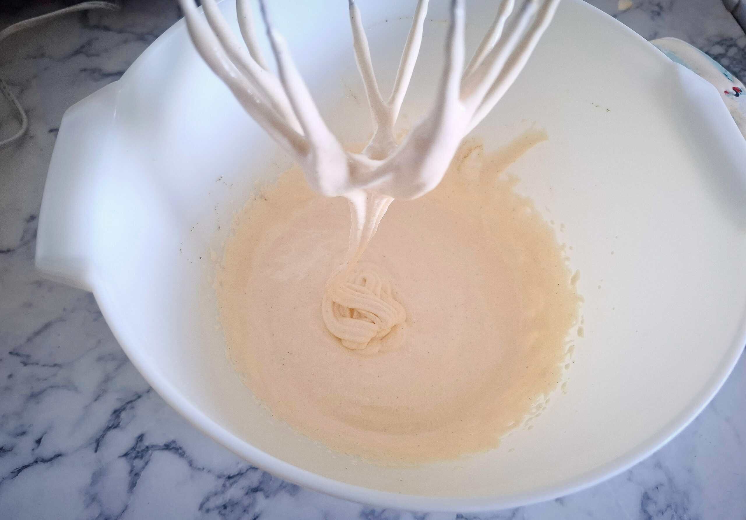 Egg yolks, sugar and vanilla have been whisked until white and fluffy in a white bowl with a whisk above it.