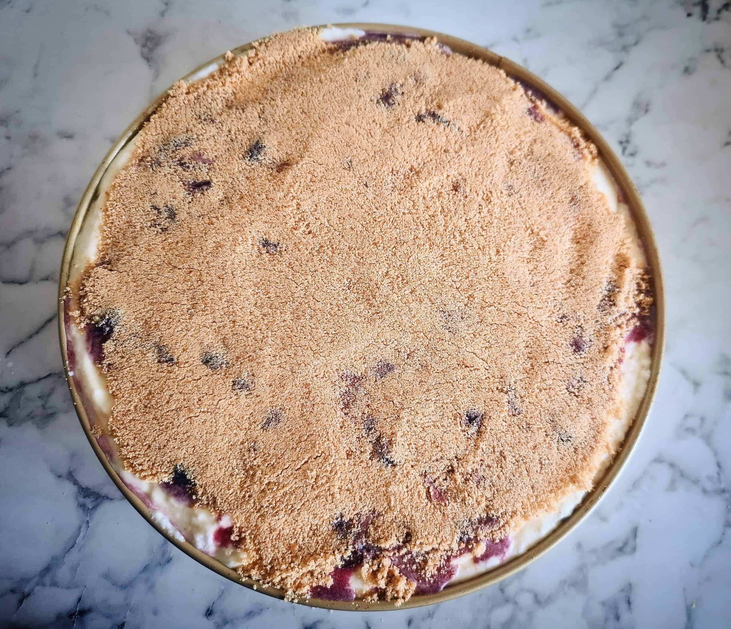 A round cake tin filled with cheese cake mixture, topped with macerated blueberries and digestive biscuit crumbs sprinkled on top.