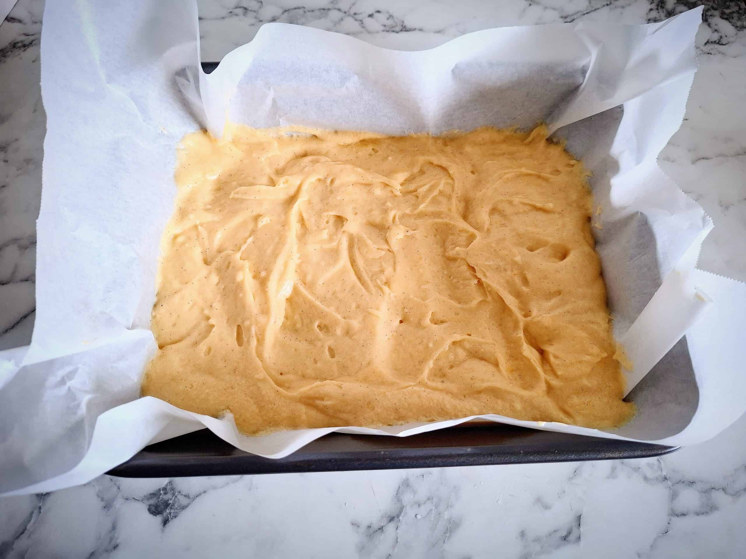 Cake batter in a black rectangular baking tin lined with parchment paper on a marbled bench top.