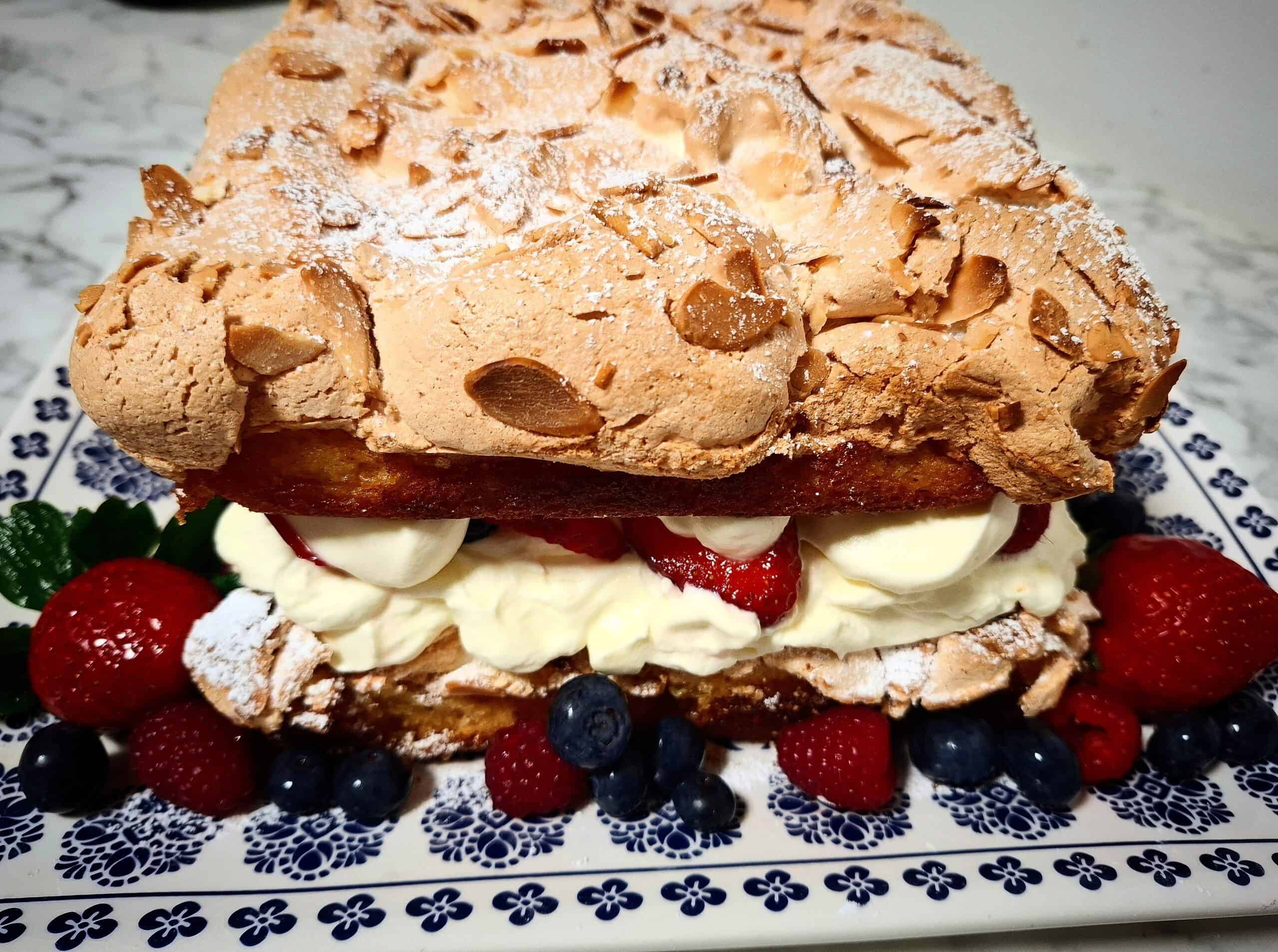 Swedish meringue cake filled with cream and fresh berries, dusted with icing sugar and decorated with fresh strawberries, blueberries and raspberries around it on a blue and white platter.