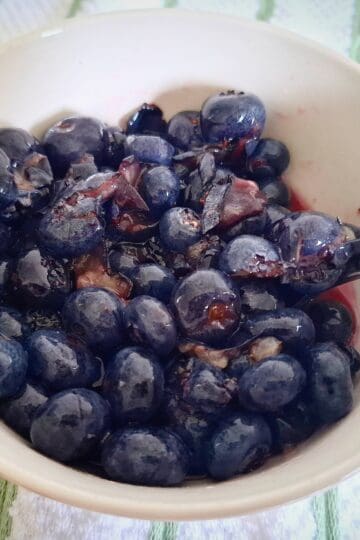 Macerated blueberries in a white, round bowl with a spoon in it on a white and green striped tea towel.
