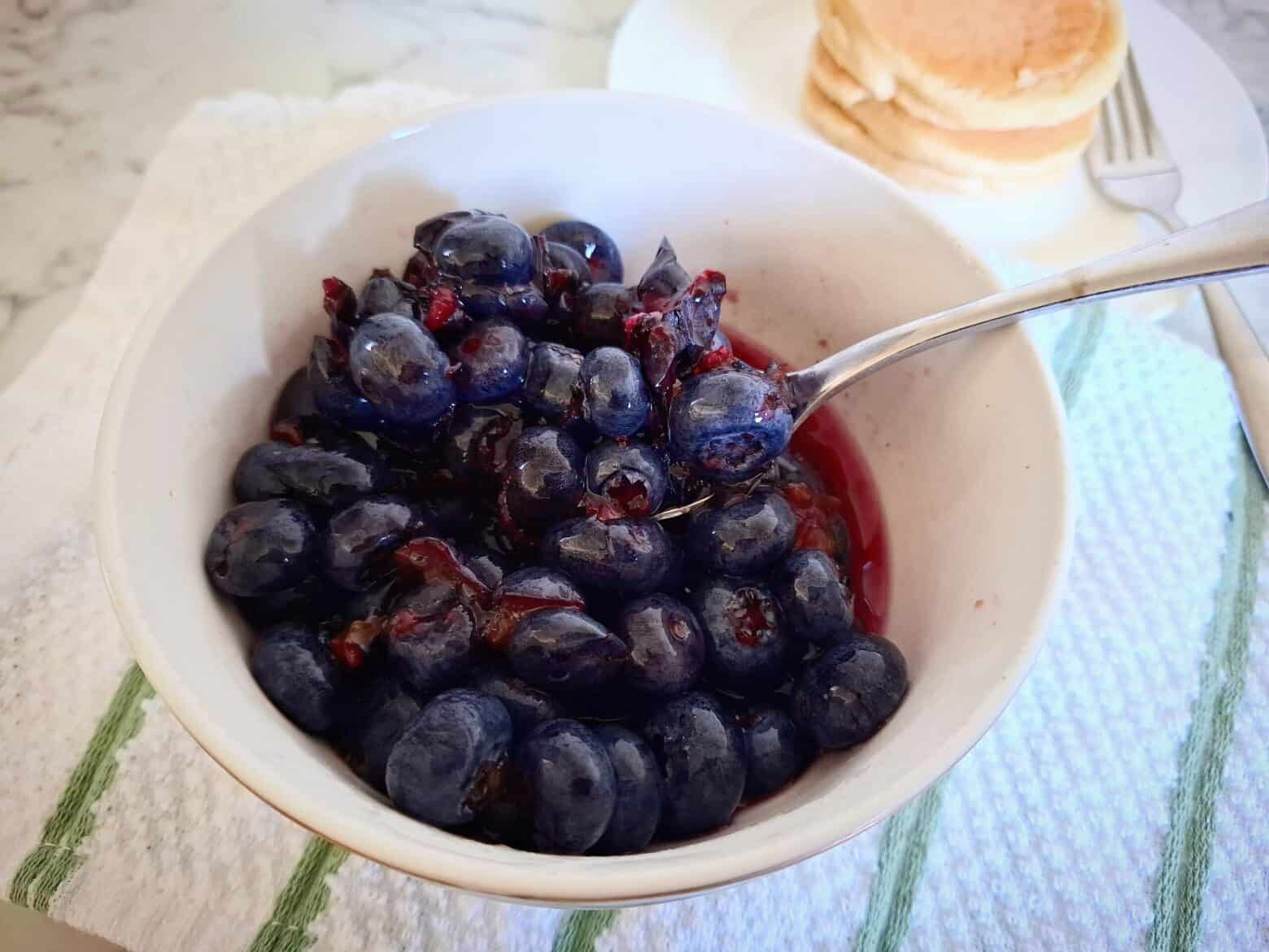 Macerated blueberries in a white, round bowl with a spoon in it on a white and green striped tea towel. A white round plate with 3 stacked pikelets and a fork behind it.
