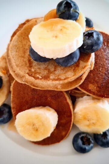Banana pikelets piled up on a white round plate with fresh blueberries and banana slice on top.