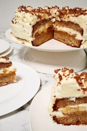 A banoffee cake with two portions cut out of it and served on two dessert plates in front of it. Two plain dessert plates and two forks behind the cake portions. A green and white striped tea towel with cake serving cutlery on top on the right hand side next to the cake.