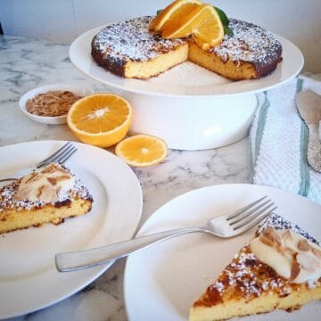 An orange and almond cake dusted with icing sugar and garnished with three swirly orange slices and a small bunch of orange leaves in the center of the cake. Two portions cut and served on two dessert plates with forks in front of the main cake. Behind the cake portions are a white ramekin of roasted almond slices and two orange halves. On the right hand side of the main cake is a green and white striped tea towel with cake serving cutlery on top.