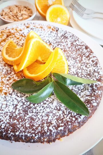 An orange and almond cake dusted with icing sugar and garnished with three swirly orange slices and a small bunch of orange leaves in the center of the cake. Behind the cake is a red ramekin of icing, a white ramekin of roasted almond slices, two orange halves and two white dessert plates with two forks on top. On the right hand side of the cake is a green and white striped tea towel with cake serving cutlery on top.