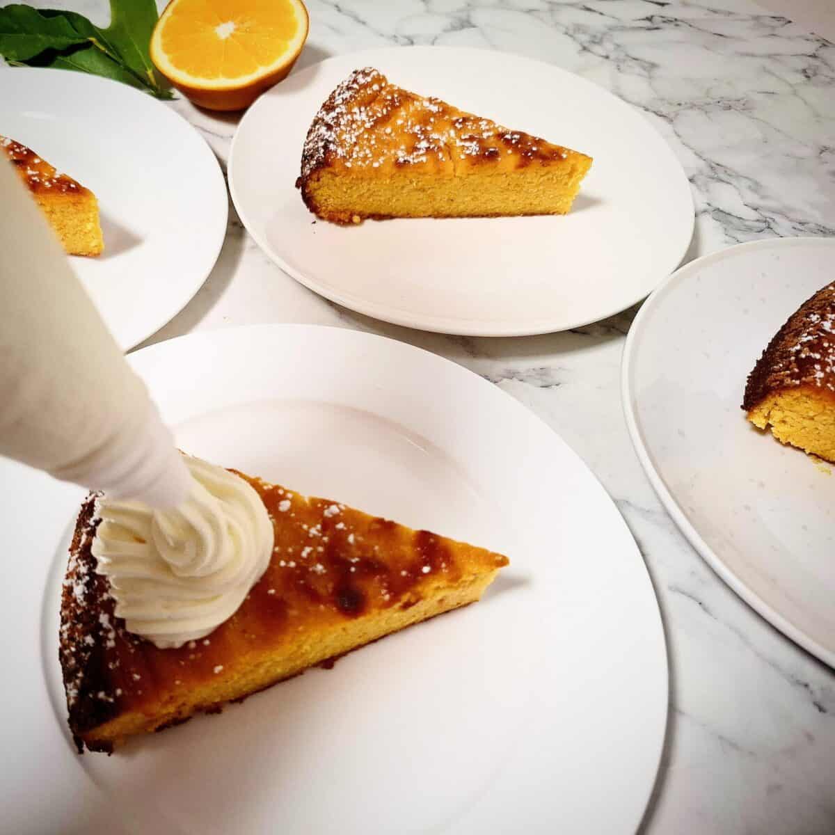 Four portions of orange and almond cake dusted with icing sugar, served on white dessert plates. Half an orange and a small bunch of orange leaves behind them. Whipped greek yogurt icing is being piped on top of one of the cake portions.