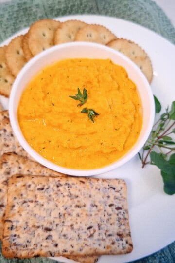 Carrot hummus served in a white ramekin topped with two thyme leaves on a white plate. On the plate are seven round crackers, five wholegrain, rectangular crackers and a bunch of fresh herbs. There is a green tea towel underneath the plate.