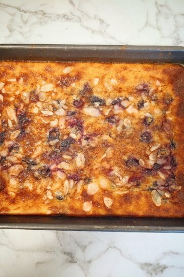 Blueberry baked custard with sliced almonds in a black rectangular baking tray on a marbled bench top.